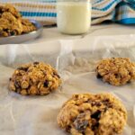 3 oatmeal raisin cookies on a parchment lined baking sheet, more cookies on a plate in the background with a glass of milk