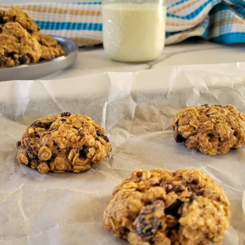 3 oatmeal raisin cookies on a parchment lined baking sheet, more cookies on a plate in the background with a glass of milk