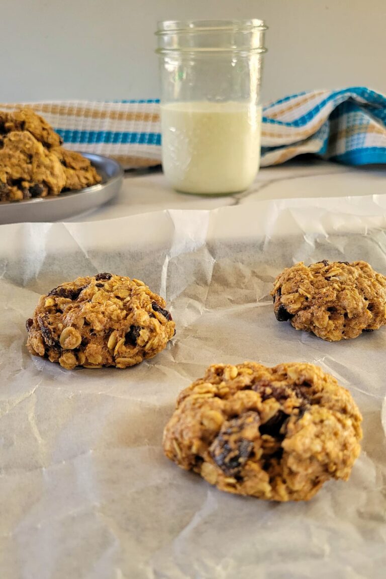 3 oatmeal raisin cookies on a parchment lined baking sheet, more cookies on a plate in the background with a glass of milk