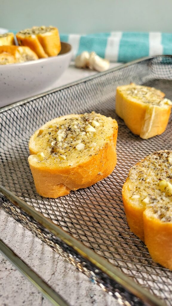 slices of garlic bread on an air fryer basket tray, bowl of more slices in the background next to a few garlic cloves