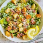 bowl of caesar salad with croutons, bacon bits, parmesan cheese, and lemon slices, next to serving spoons and a bowl of bacon bits