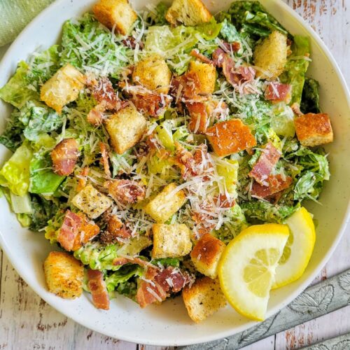 bowl of caesar salad with croutons, bacon bits, parmesan cheese, and lemon slices, next to serving spoons and a bowl of bacon bits