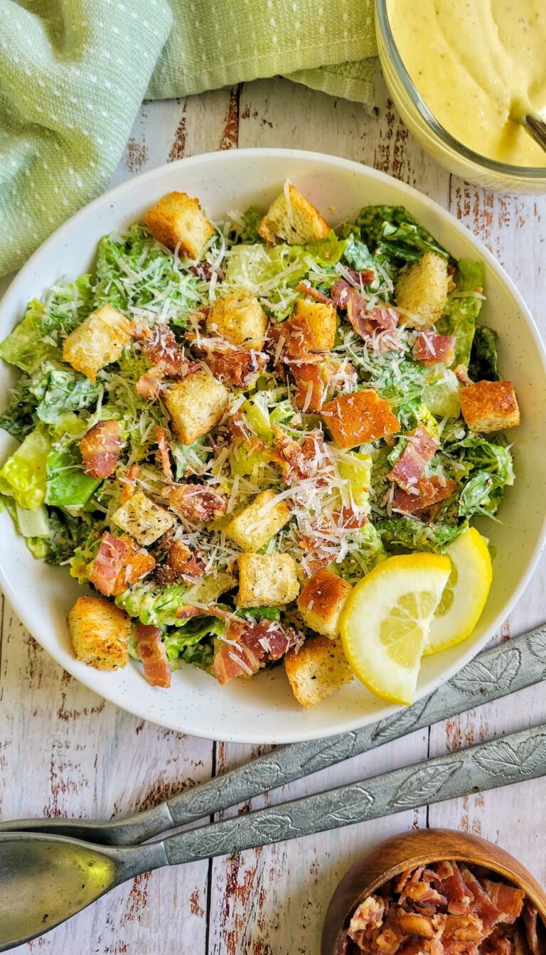 bowl of caesar salad with croutons, bacon bits, parmesan cheese, and lemon slices, next to serving spoons and a bowl of bacon bits