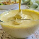 bowl of caesar salad dressing with a spoon lifting out of it, caesar salad in the background