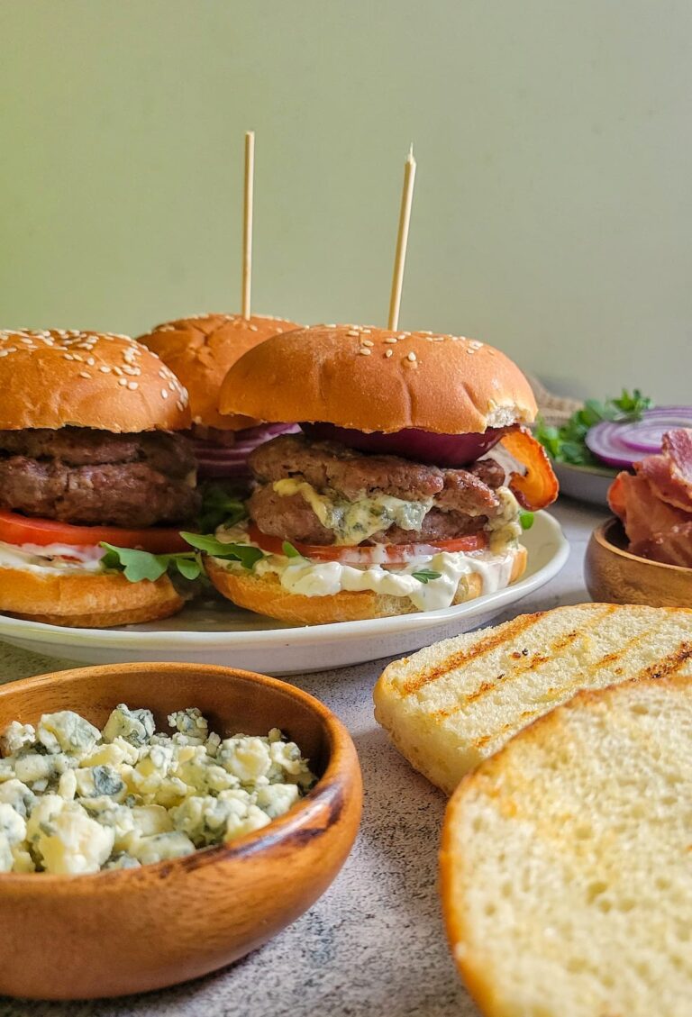 3 burgers on buns with toppings on a plate, bowl of blue cheese next to grilled burger buns in the front