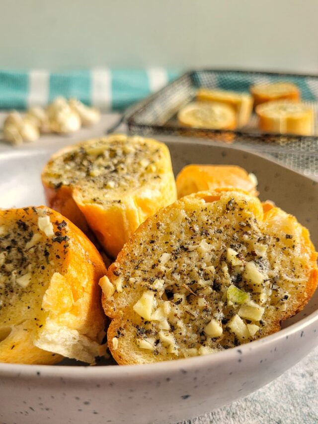 bowl of sliced garlic bread, air fryer tray in the background with more slices next to some unpeeled garlic cloves
