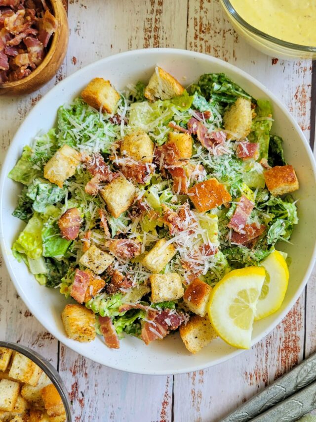 caesar salad with croutons, bacon, parmesan cheese and lemon slices in a bowl, small bowls of croutons, bacon bits and dressing in the background