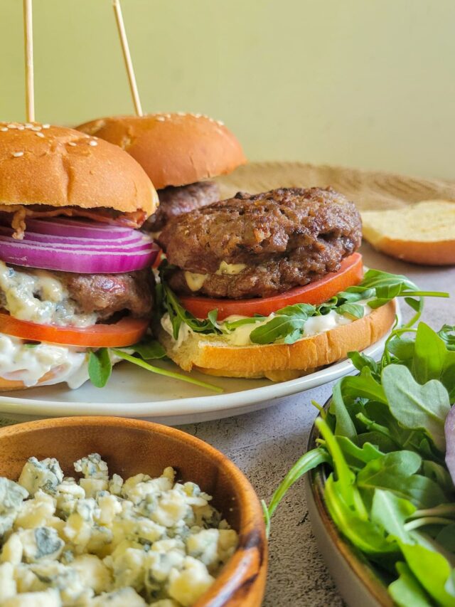 blue cheese stuffed burgers on buns on a plate with toppings, bowl of blue cheese and more toppings in the front