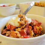 bowl of strawberry rhubarb crisp with whipped cream and a spoon, more in the background