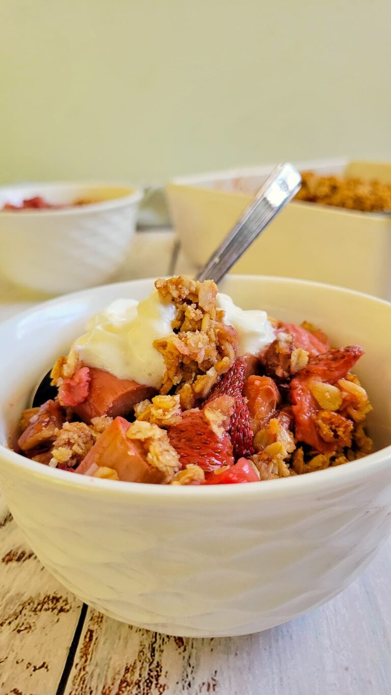 bowl of strawberry rhubarb crisp with whipped cream and a spoon, more in the background