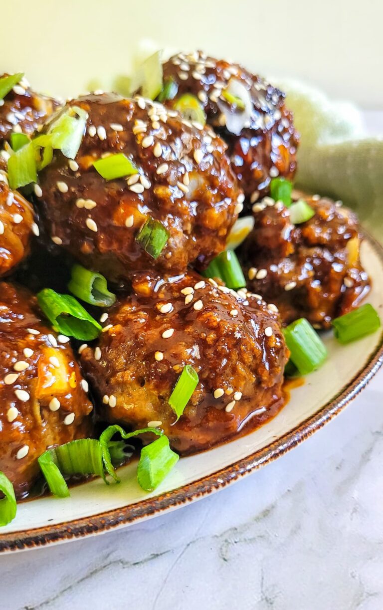 sticky asian meatballs in a brown sauce on a plate with sesame seeds and green onions.