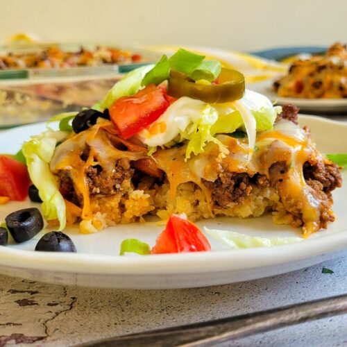piece of taco casserole topped with jalapenos, black olives, tomatoes, sour cream, lettuce, and green onions on a plate, more in the background