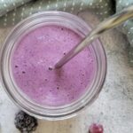 purple smoothie in a glass with a metal straw surrounded by berries