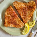 two cooked seasoned tuna steaks on a plate with two slices of lemon, bowl of greens in the background