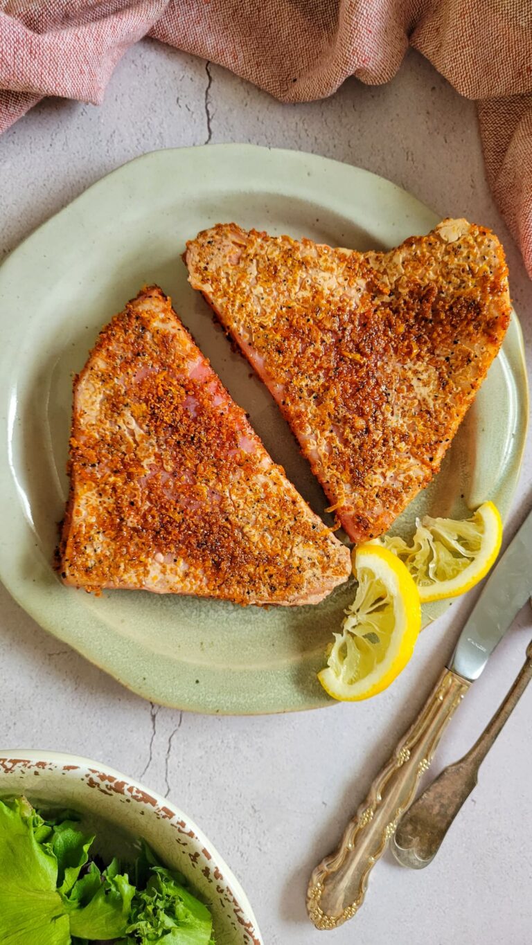 two cooked seasoned tuna steaks on a plate with two slices of lemon, bowl of greens in the background