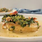 creamy pork chop on a plate topped with bacon and spinach, another one in the background