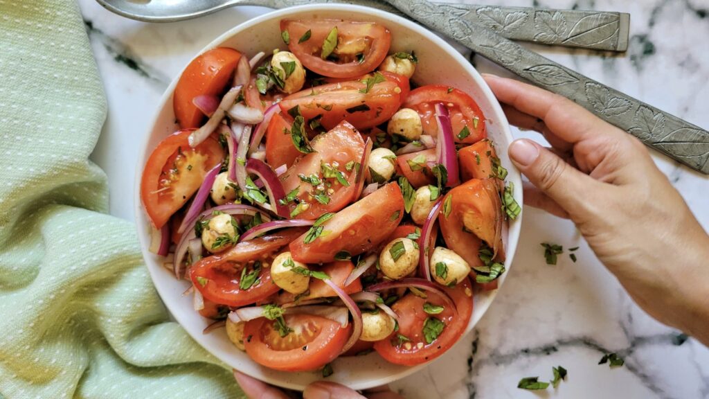 hand holding a bowl of tomato salad with fresh basil, mozzarella balls, red onions, and a balsamic dressing