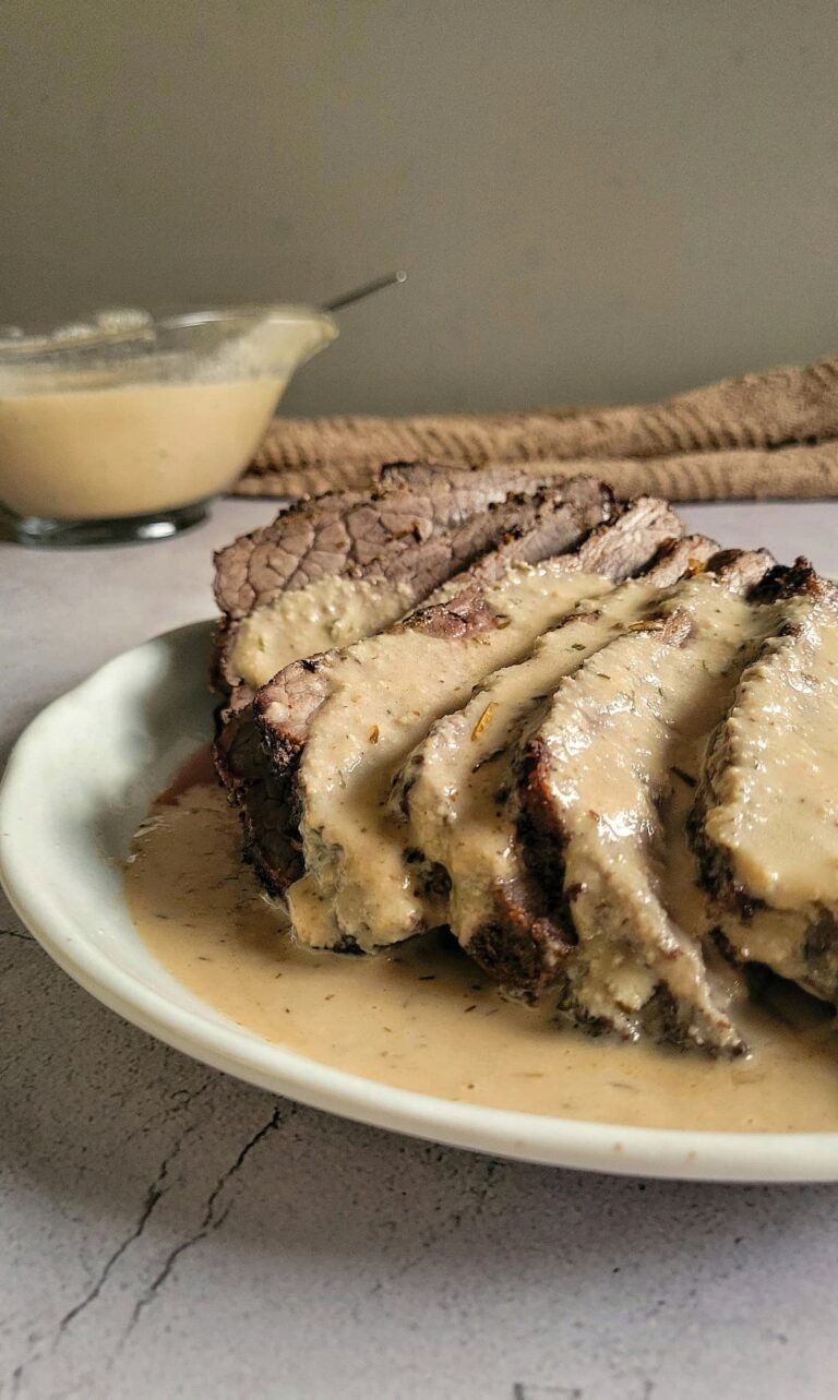 sliced beef on a plate with pale brown gravy over top. Gravy boat with more in the background