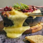 baked brie spilling out of a cast iron skillet, rosemary, cranberries and pecans on top, crackers in the front