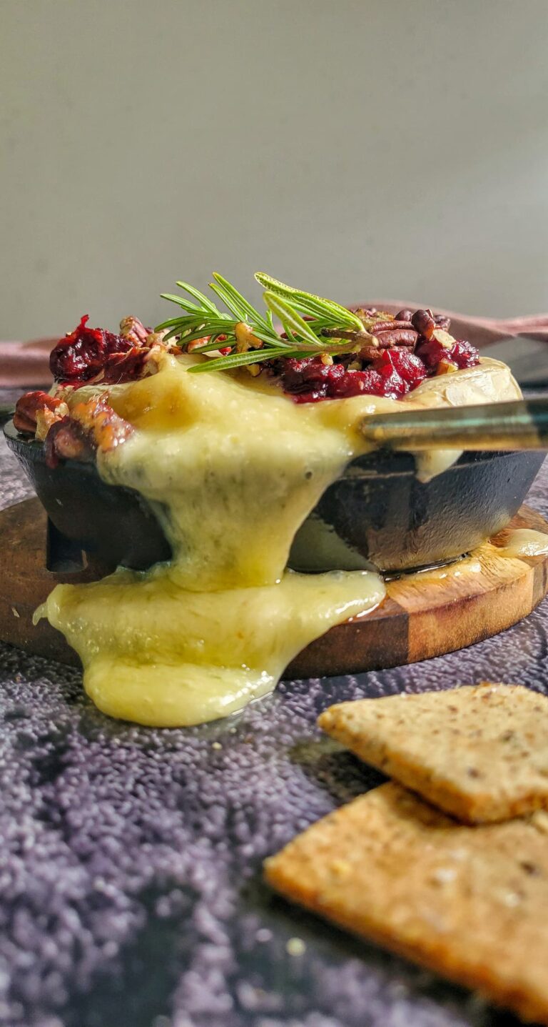 baked brie spilling out of a cast iron skillet, rosemary, cranberries and pecans on top, crackers in the front