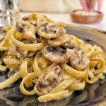 fettuccine mushroom alfredo stacked on a plate, hand grabbing parm cheese out of a ramekin in the background next to a pepper grinder.
