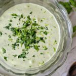 cilantro lime crema topped with chopped cilantro in a bowl, next to a spoon, wedge of lime, and more cilantro