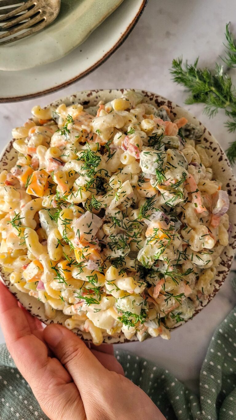 hand holding a bowl of dill pickle pasta salad with chopped fresh dill, pickles, cubed cheddar cheese, and onions