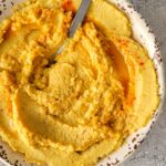 hummus sprinkled with paprika and olive oil in a bowl with a spoon, crackers and garlic around it.