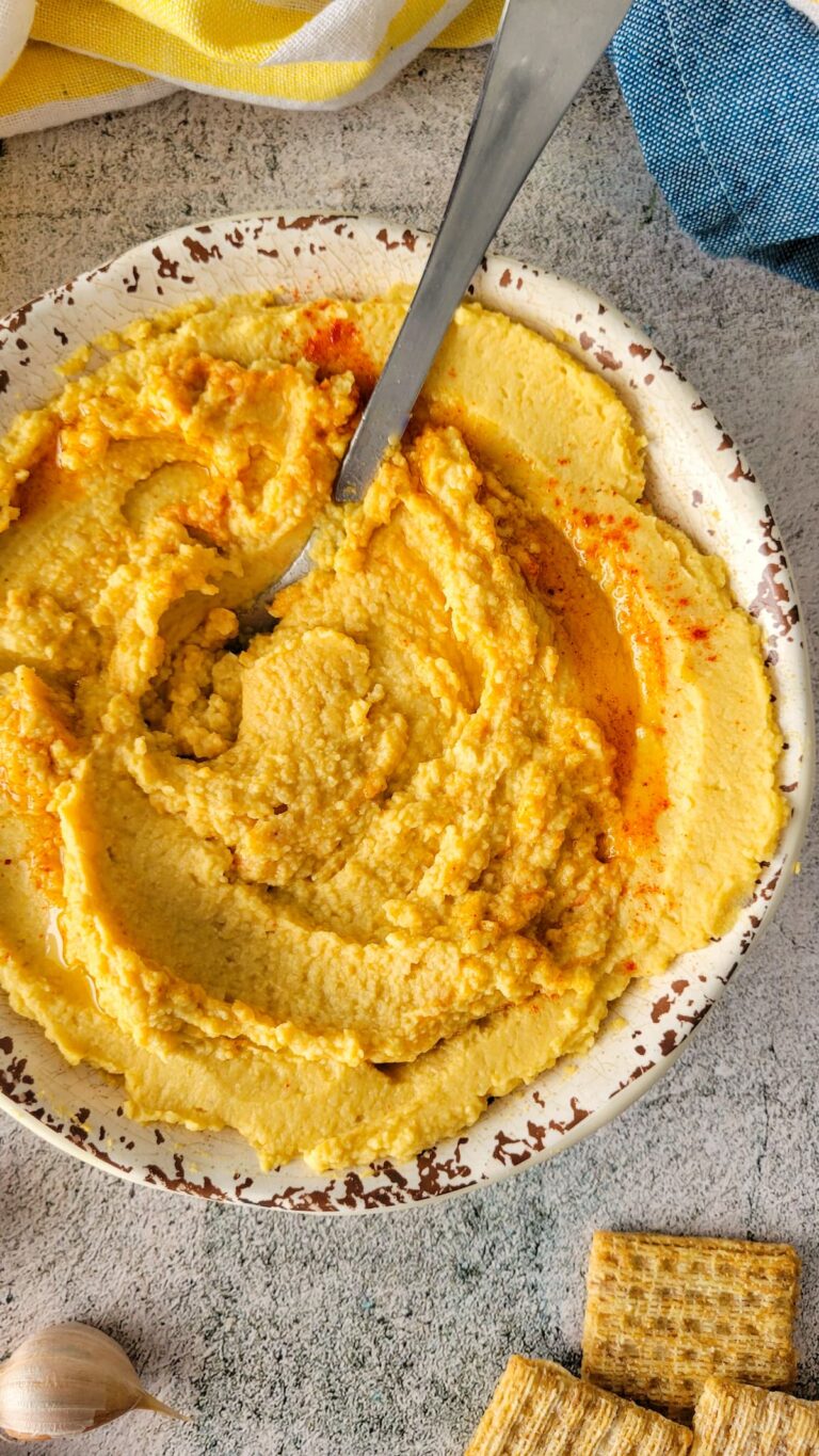 hummus sprinkled with paprika and olive oil in a bowl with a spoon, crackers and garlic around it.