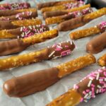 milk chocolate covered pretzel rods with sprinkles on a parchment lined baking sheet