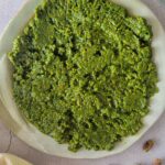 pesto on a plate next to a bowl of dry pasta, garlic and pistachios around