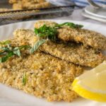 3 chicken cutlets on a plate with basil and lemon, more on a tray in the background