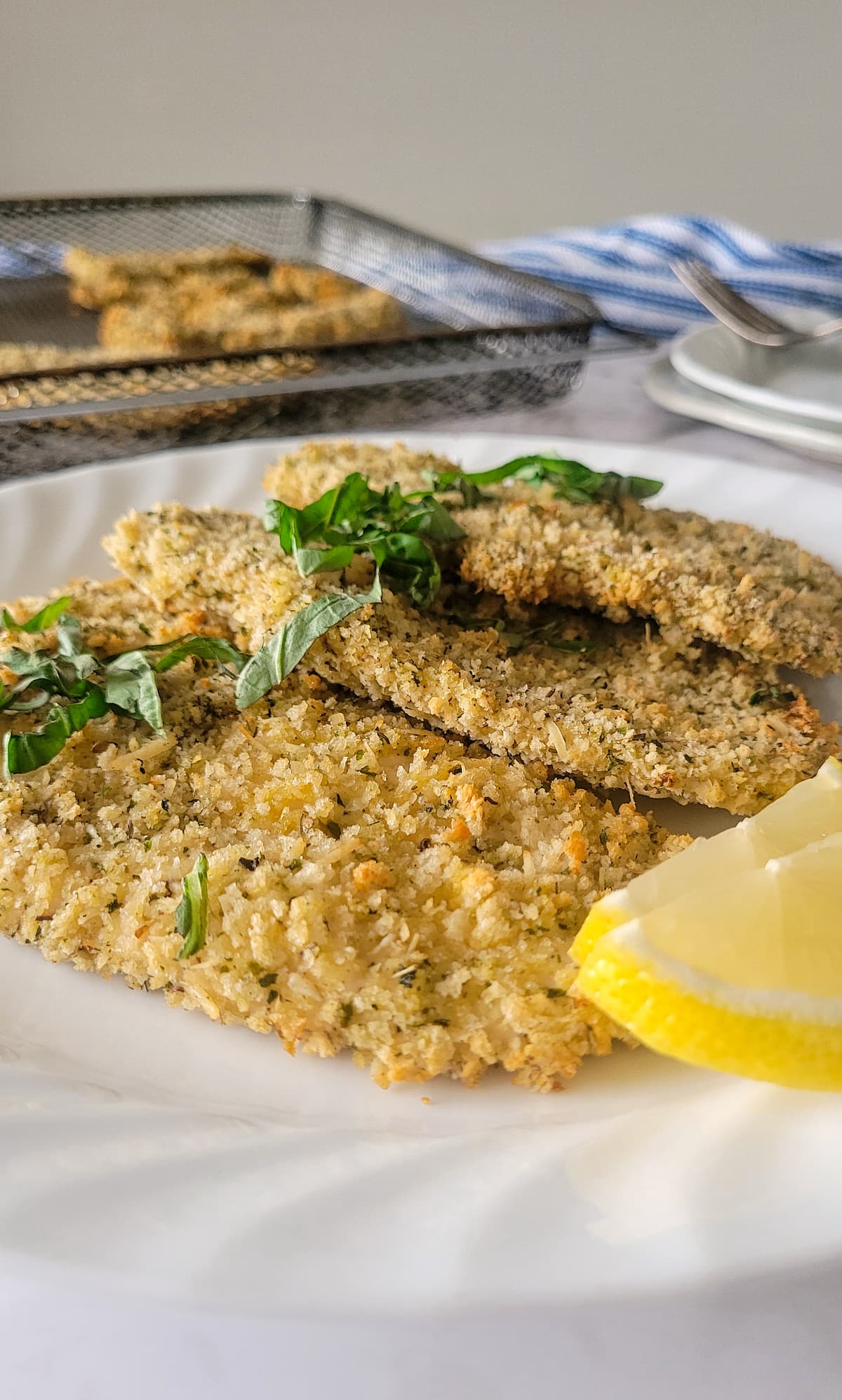 3 chicken cutlets on a plate with basil and lemon, more on a tray in the background
