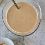 bowl of creamy balsamic dressing with a spoon, bowl of lettuce in the background along with a white plate with a mini whisk.