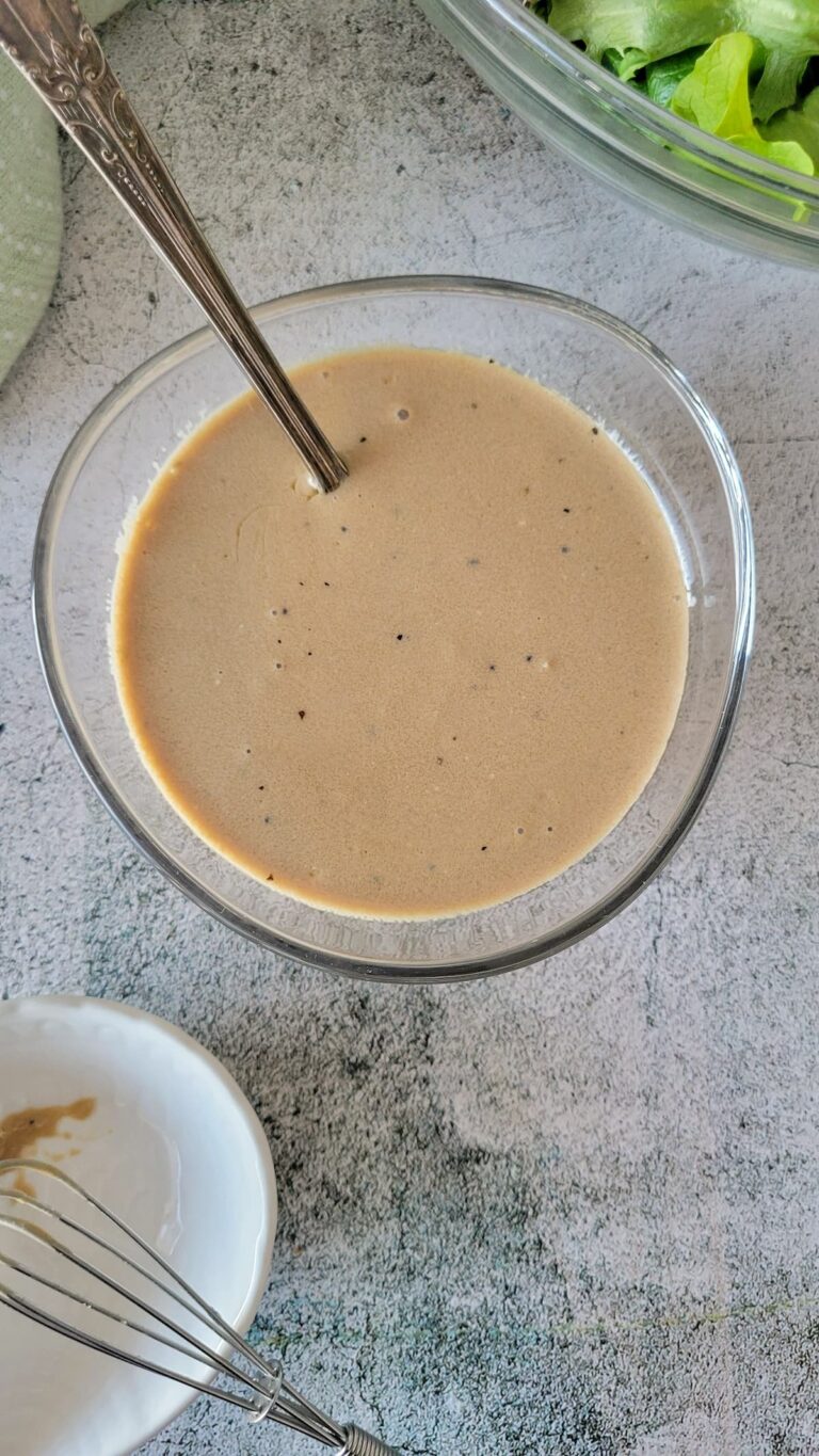 bowl of creamy balsamic dressing with a spoon, bowl of lettuce in the background along with a white plate with a mini whisk.