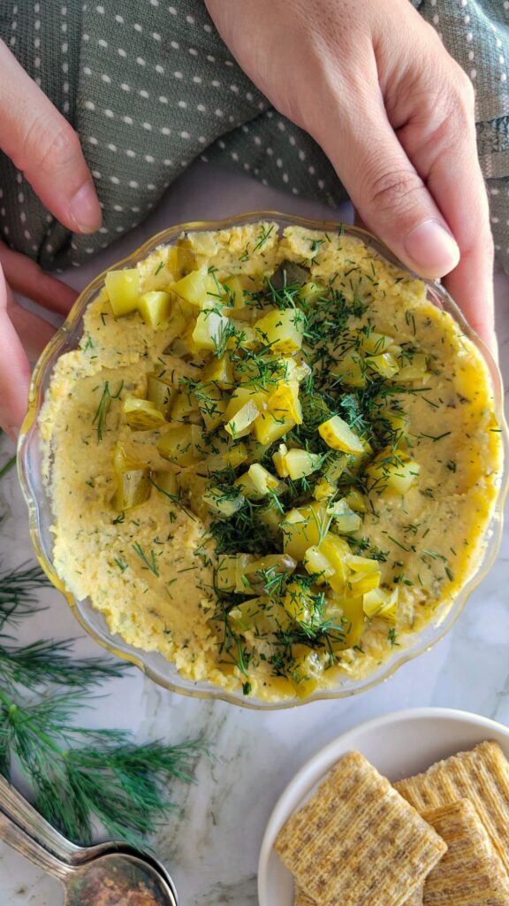 hand holding a bowl of hummus with chopped pickles and dill, crackers next to it