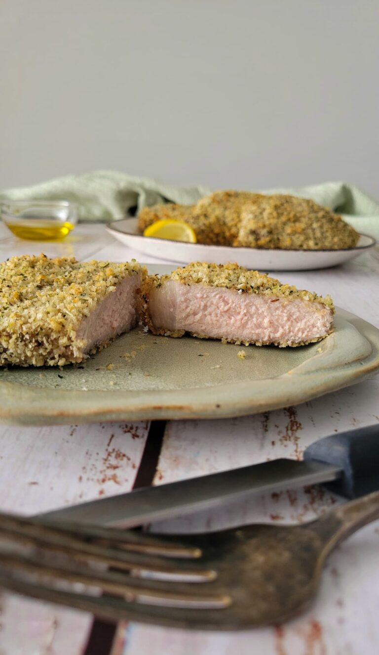 thick breaded pork chop on a plate, cut in half, fork and knife in the front, more chops in the background