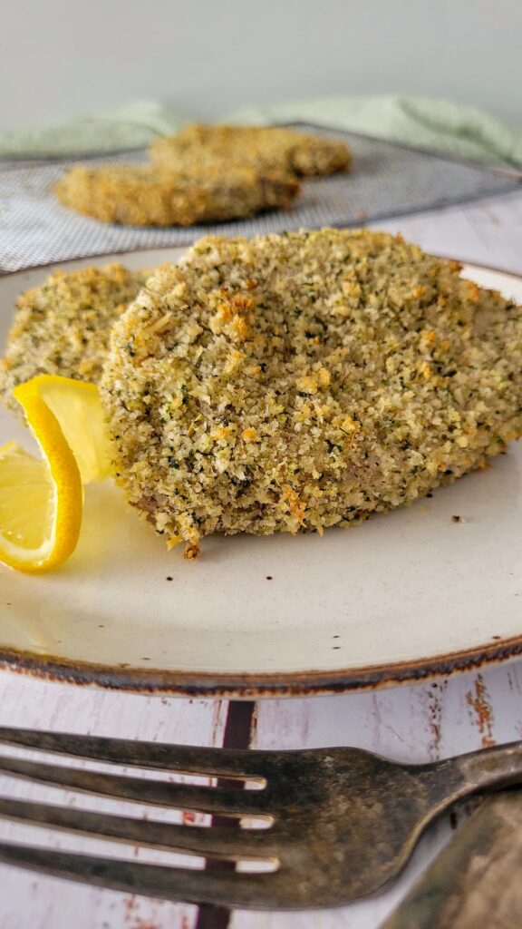 two breaded pork chops on a plate with lemon, more on an air fryer tray in the background, fork in the front