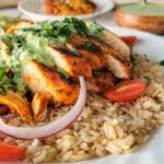 sliced chicken breast on a plate with brown rice, cherry tomatoes, red onions, green sauce, and cilantro, more green sauce and chicken in the background