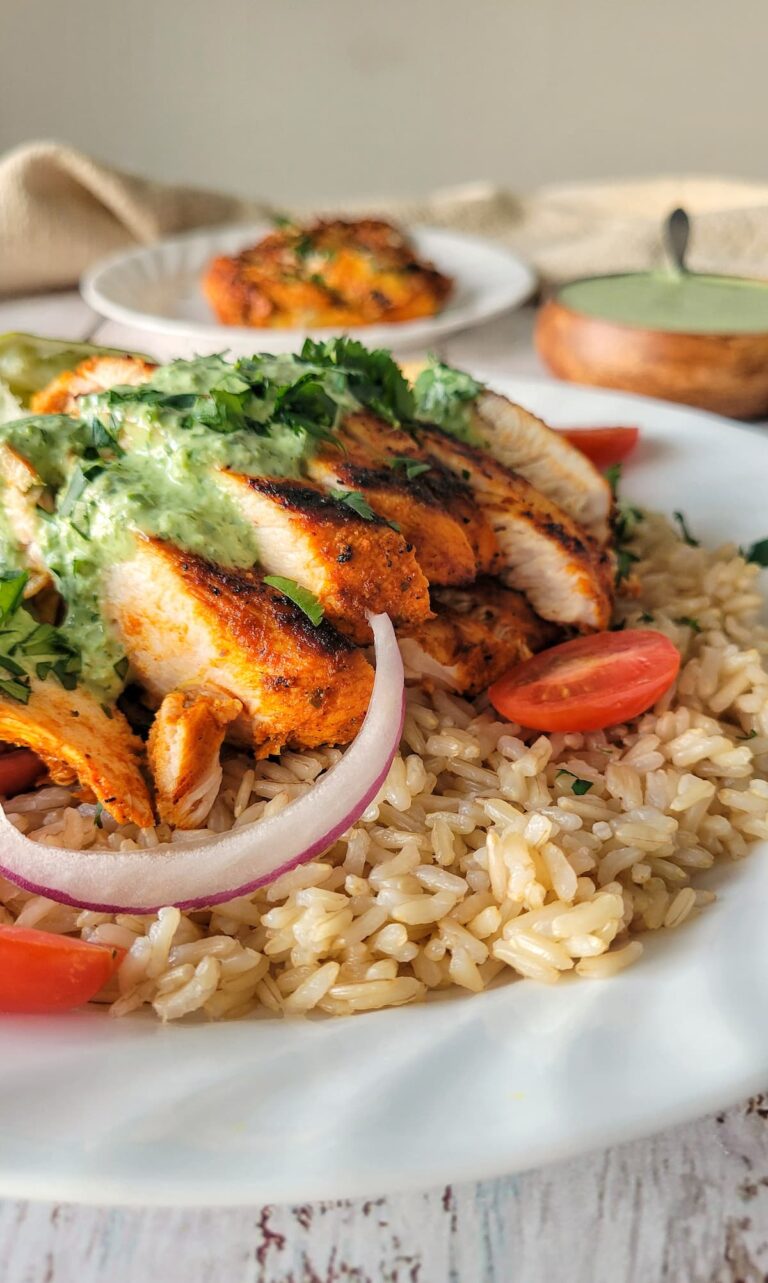 sliced chicken breast on a plate with brown rice, cherry tomatoes, red onions, green sauce, and cilantro, more green sauce and chicken in the background