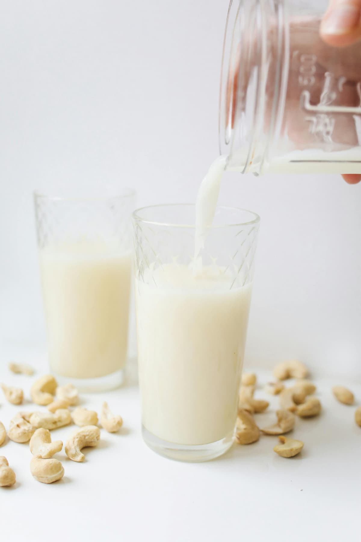 milk being poured into a glass, another glass behind it, cashews around it