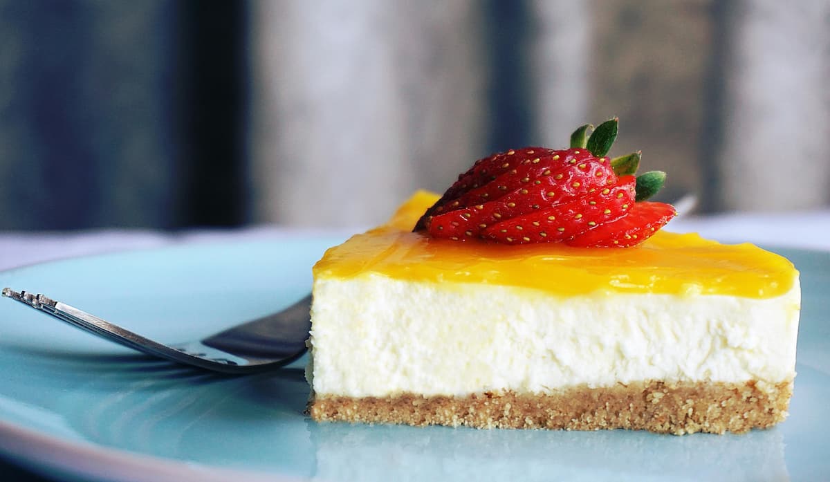 slice of cheesecake with graham cracker crust, yellow topping and a sliced strawberry