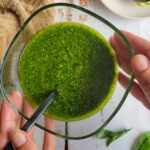 hands holding a bowl of pesto with a spoon in it, garlic cloves, basil leaves and lemon in the background