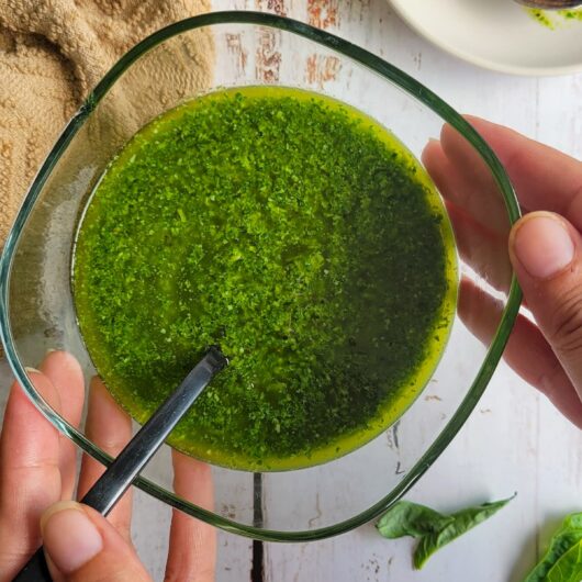 hands holding a bowl of pesto with a spoon in it, garlic cloves, basil leaves and lemon in the background