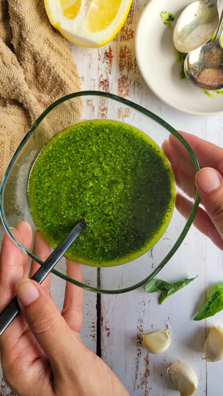 hands holding a bowl of pesto with a spoon in it, garlic cloves, basil leaves and lemon in the background