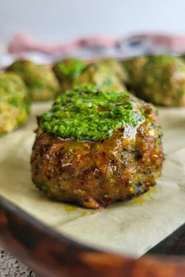 cooked meatball on a baking sheet topped with pesto, more in the background