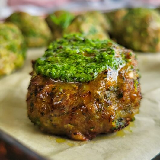cooked meatball on a baking sheet topped with pesto, more in the background