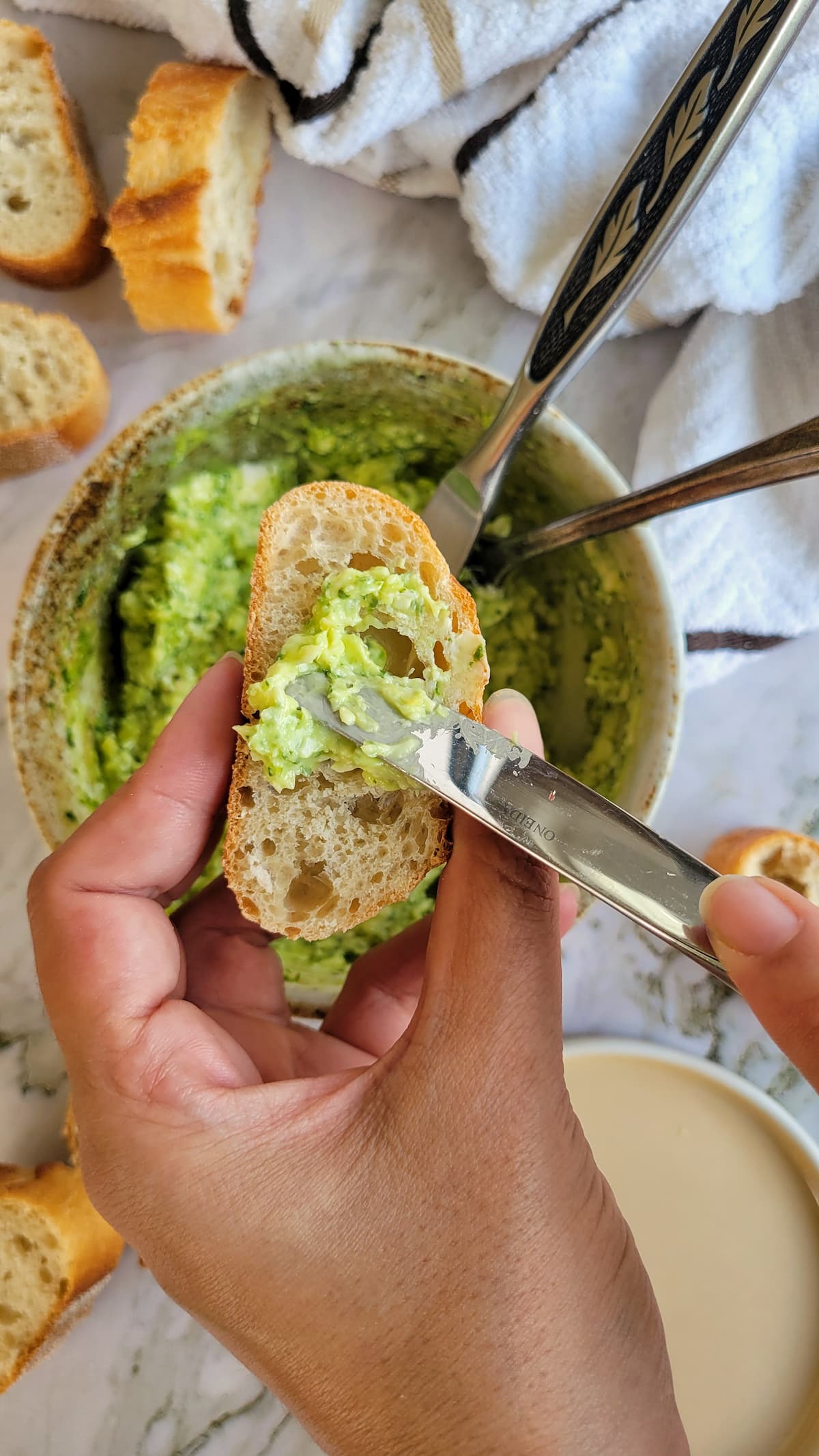 hand with a knife spreading pesto butter on a slice of bread over a bowl with more