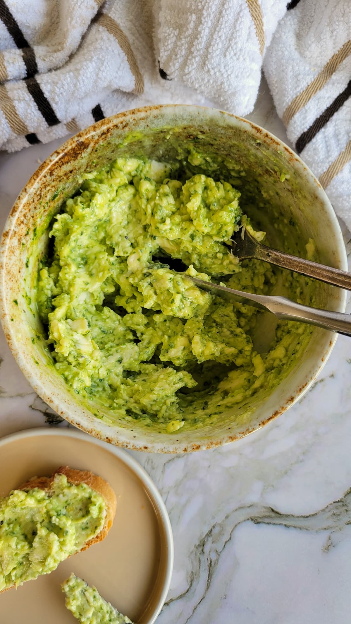 bowl of pesto butter with utensils next to a plate of bread smeared with some of the butter
