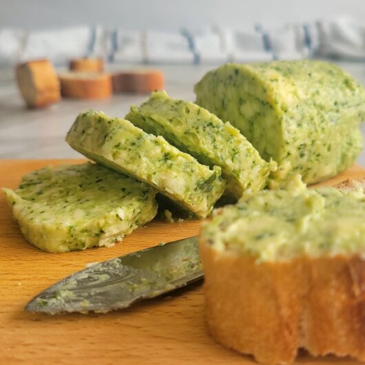 log of pesto butter on a cutting board, some cut into slices, bread in the front and back